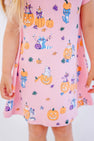 A child wearing The Beaufort Bonnet Company’s Polly Play Dress - Candy & Critters (Sandpearl Pink), made from soft Pima Cotton and decorated with whimsical pumpkins, raccoons, and leaves. Only the lower half of her dress is visible, showing light skin and partially revealing her long blonde hair.