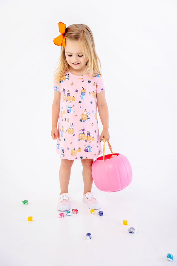 A young girl with a bright orange bow in her hair wears the charming Polly Play Dress - Candy & Critters (Sandpearl Pink) by The Beaufort Bonnet Company, crafted from soft Pima Cotton and adorned with whimsical pumpkins and cats. She holds a pink pumpkin bucket and stands among scattered colorful candies on a white background, making this girls’ dress perfect for festive occasions.