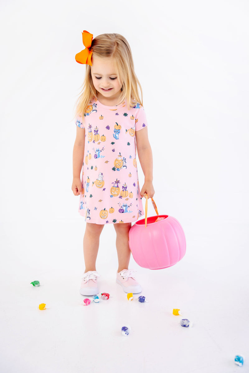 A young girl with a bright orange bow in her hair wears the charming Polly Play Dress - Candy & Critters (Sandpearl Pink) by The Beaufort Bonnet Company, crafted from soft Pima Cotton and adorned with whimsical pumpkins and cats. She holds a pink pumpkin bucket and stands among scattered colorful candies on a white background, making this girls’ dress perfect for festive occasions.