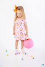 A young girl with a bright orange bow in her hair wears the charming Polly Play Dress - Candy & Critters (Sandpearl Pink) by The Beaufort Bonnet Company, crafted from soft Pima Cotton and adorned with whimsical pumpkins and cats. She holds a pink pumpkin bucket and stands among scattered colorful candies on a white background, making this girls’ dress perfect for festive occasions.