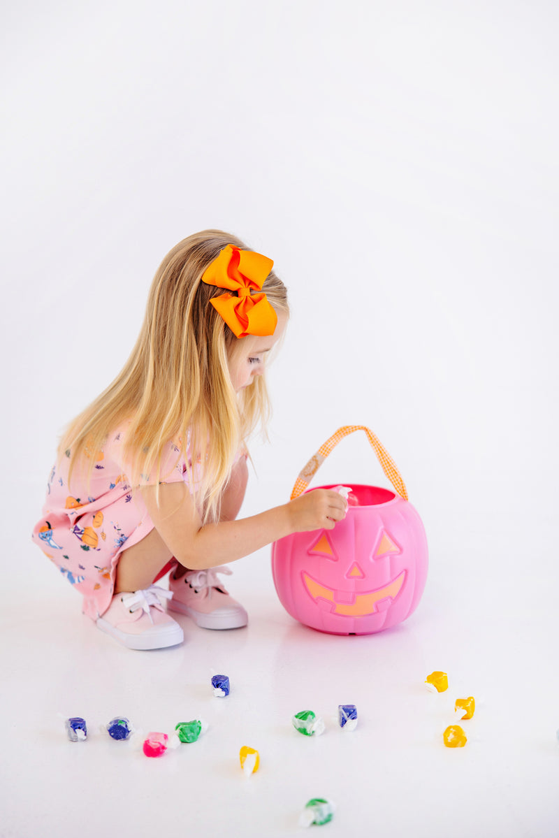 A young girl wearing The Beaufort Bonnet Company’s Polly Play Dress - Candy & Critters (Sandpearl Pink), adorned with a large orange bow, gazes into a pink plastic pumpkin bucket as wrapped candies are scattered around her on a white floor.