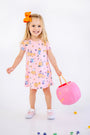 Here’s your rewritten sentence using the provided product data: A smiling young girl with blond hair tied in an orange bow holds a pink pumpkin bucket. She wears the Polly Play Dress - Candy & Critters (Sandpearl Pink) by The Beaufort Bonnet Company, paired with matching pink shoes. Crafted from soft Pima Cotton for extra comfort, this girls dress stands out as colorful candies are scattered across the white floor around her.
