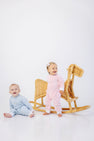 Two babies in pastel pajamas pose near a wicker rocking horse. One baby sits on the floor in a blue playsuit, while the other stands beside the rocking horse wearing The Beaufort Bonnet Company’s Paxton Playsuit in Palm Beach Pink with Rocking Horse Embroidery and a white bow headband. The background is white and minimal.