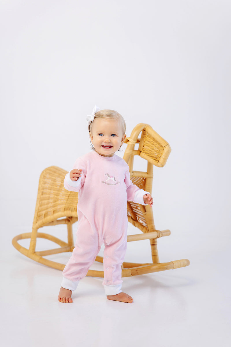 A toddler smiles in front of a light-colored wicker rocking horse, wearing The Beaufort Bonnet Company’s Paxton Playsuit in Palm Beach Pink with Rocking Horse Embroidery and white cuffs, her hair adorned with a white bow against a plain white background.
