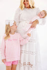 A young girl wearing a Cassidy Comfy Crewneck - Palm Beach Pink Gingham with Hamptons Hot Pink Big Sister looks up at her mother, who is holding a newborn baby in pink footed pajamas.