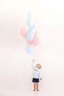 A young boy wears a Cassidy Comfy Crewneck - Buckhead Blue Gingham with Nantucket Navy Big Brother shirt while holding a large bunch of blue and pink balloons for a sibling reveal.