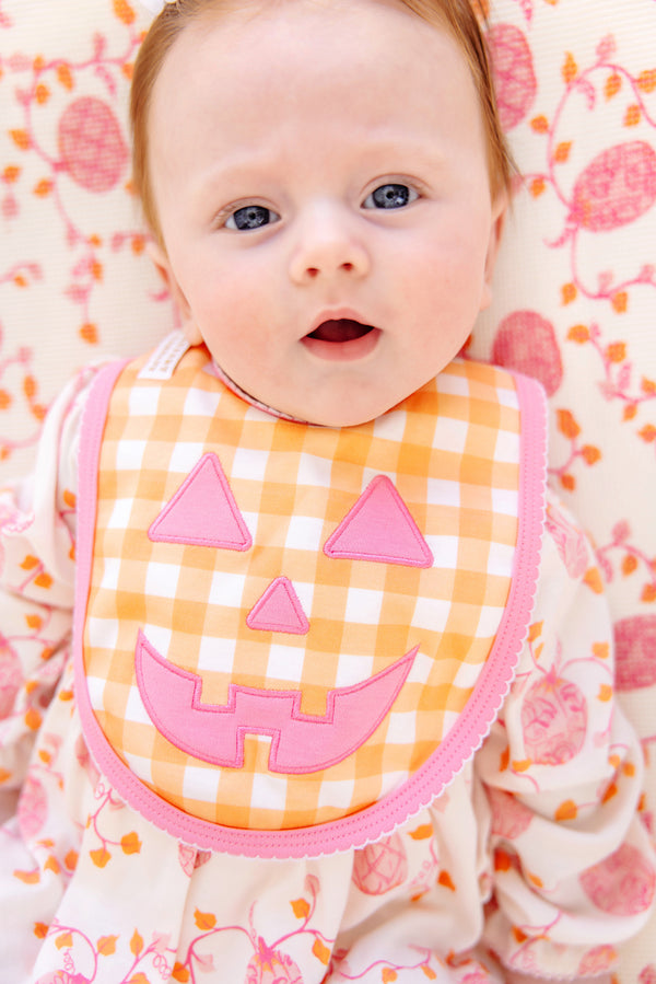 A baby with blue eyes lies on a patterned blanket, wearing a white and pink outfit and an orange Burp Me Bib - Seashore Sherbet Gingham & Hamptons Hot Pink from The Beaufort Bonnet Company, perfect as a fall baby essential.