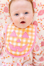 A baby with blue eyes lies on a patterned blanket, wearing a white and pink outfit and an orange Burp Me Bib - Seashore Sherbet Gingham & Hamptons Hot Pink from The Beaufort Bonnet Company, perfect as a fall baby essential.