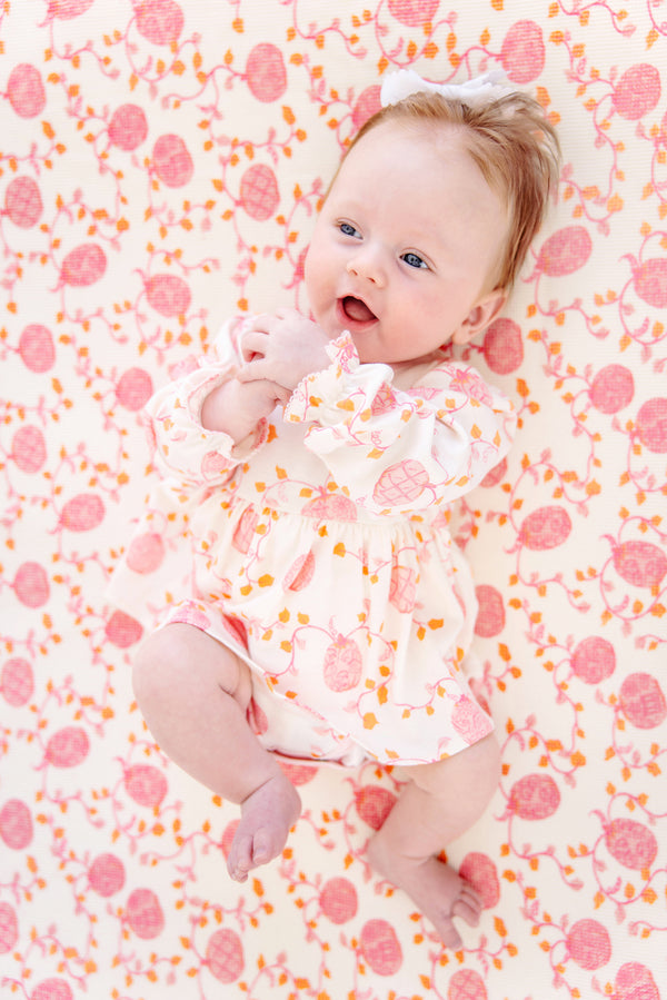 A baby with light skin and reddish hair lies on a cozy Silent Night Throw - Gourd-geous (Palmetto Pearl) with Romany Rose by The Beaufort Bonnet Company, wearing a matching dress and bow. The baby is smiling, looking up, and holding their hands together. Both the blanket and dress feature charming pink and orange designs.