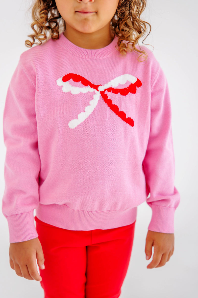 Child wearing Isabelle’s Intarsia Sweater - Hamptons Hot Pink with Scalloped Bow, featuring a pink sweater with a red and white scalloped bow design on the chest.
