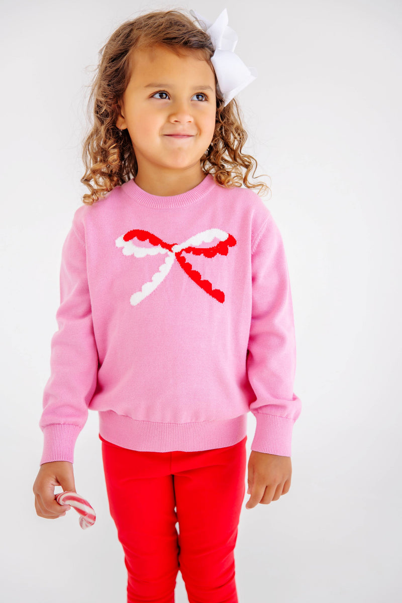 A young girl wearing a pink sweater with a red and white bow design and Mitzy Sue Slacks - Richmond Red, holding a candy cane.