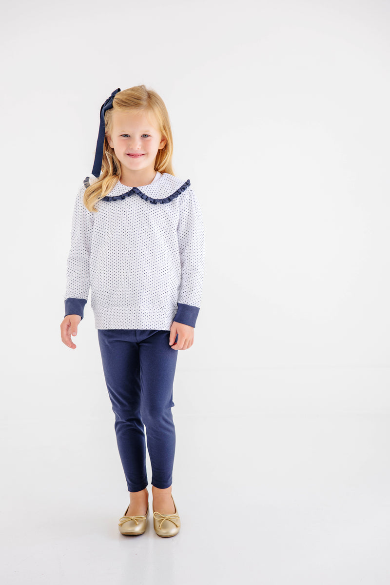 A young child with blonde hair stands and smiles against a plain white background, wearing The Beaufort Bonnet Company’s Maude’s Ruffle Cassidy Comfy Crewneck in Nantucket Navy Microdot with Worth Avenue White. She pairs the top with navy leggings, gold shoes, and a matching navy bow in her hair.