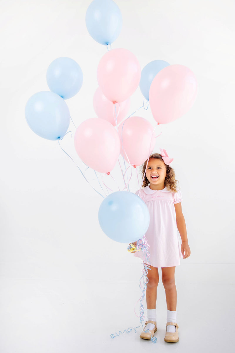 A smiling girl in a Holly Day Dress - Palm Beach Pink with Hamptons Hot Pink Big Sister holds a large cluster of pink and light blue balloons.