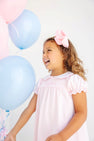 A young girl wears a Holly Day Dress - Palm Beach Pink with Hamptons Hot Pink Big Sister with a matching hair bow and stands next to a cluster of pink and light blue balloons.