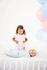 A young girl in a pink dress looks down into a crib at a baby wearing a Cameron Cross Front Set - Buckhead Blue with Nantucket Navy Little Brother, accompanied by blue and pink balloons for this baby announcement.