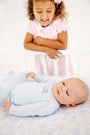 A young girl smiles as she looks down at her little brother, who is wearing a Cameron Cross Front Set - Buckhead Blue with Nantucket Navy Little Brother onesie while lying in a crib.