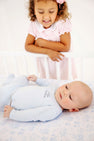 A young girl smiles as she looks down at her little brother, who is wearing a Cameron Cross Front Set - Buckhead Blue with Nantucket Navy Little Brother onesie while lying in a crib.