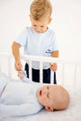 A young boy in a Carter Crewneck - Buckhead Blue with Nantucket Navy Big Brother holds the hand of an infant lying in a white crib.