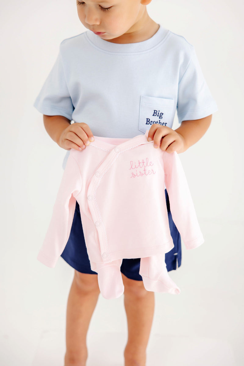 A young child wearing a Buckhead Blue with Nantucket Navy Big Brother Carter Crewneck holds up a pale pink long-sleeved infant onesie embroidered with little sister.