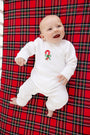 A smiling baby in The Beaufort Bonnet Company’s Paxton Playsuit – Palmetto Pearl with Candy Cane, a white velour knit holiday baby outfit, lying on a red plaid blanket.