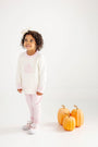 A young girl wearing The Beaufort Bonnet Company’s Cassidy Comfy Crewneck in Palmetto Pearl with Palm Beach Pink & Pumpkin Applique stands smiling beside three orange pumpkins on a white floor against a plain white background.