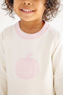 A smiling child with curly hair wears The Beaufort Bonnet Company’s Cassidy Comfy Crewneck—a cozy girls’ fall sweatshirt in Palmetto Pearl, featuring a Palm Beach Pink collar and a charming pink gingham pumpkin applique on the front.