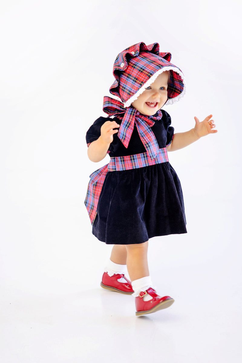 A smiling toddler walks against a white background, wearing The Beaufort Bonnet Company’s Bellefaire Bonnet in Bucktown Tartan with Worth Avenue White Eyelet, paired with a navy dress featuring red Bucktown Tartan accents and red shoes.