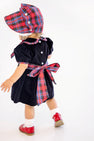A toddler models the Bellefaire Bonnet - Bucktown Tartan with Worth Avenue White Eyelet by The Beaufort Bonnet Company, paired with a velvet dress featuring a red Bucktown Tartan sash, white socks, and red shoes, standing and facing away on a white background.