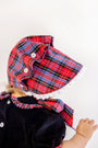 A baby is shown from behind against a plain white background, wearing The Beaufort Bonnet Company’s Bellefaire Bonnet in Bucktown Tartan with Worth Avenue White Eyelet, paired with a velvet outfit featuring matching tartan accents.