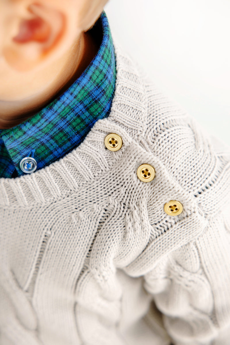 Close-up of a child wearing a cream cable-knit sweater with three tan buttons on the shoulder, layered over The Beaufort Bonnet Company’s Dean’s List Dress Shirt in Woodlake Point Plaid. Only the lower face, ear, and shoulder are visible.