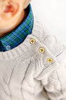 Close-up of a child wearing a cream cable-knit sweater with three tan buttons on the shoulder, layered over The Beaufort Bonnet Company’s Dean’s List Dress Shirt in Woodlake Point Plaid. Only the lower face, ear, and shoulder are visible.