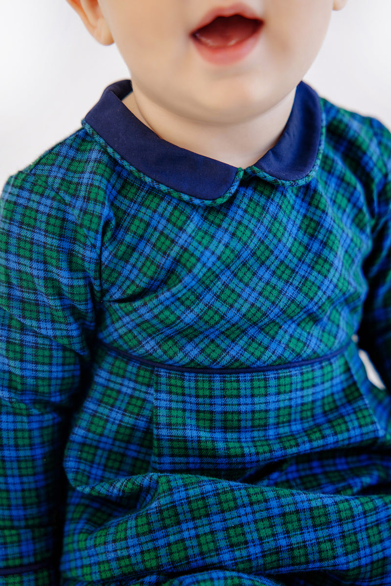 A young child wearing The Beaufort Bonnet Company’s Long Sleeve Bradford Romper in Woodlake Point Plaid with Nantucket Navy, smiling with their mouth slightly open. The image is cropped to show only the child’s lower face and upper body.