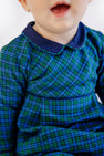 A young child wearing The Beaufort Bonnet Company’s Long Sleeve Bradford Romper in Woodlake Point Plaid with Nantucket Navy, smiling with their mouth slightly open. The image is cropped to show only the child’s lower face and upper body.