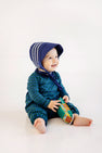 A smiling baby in a blue bonnet and The Beaufort Bonnet Company’s Long Sleeve Bradford Romper – Woodlake Point Plaid with Nantucket Navy sits on the floor, holding a colorful toy bus.