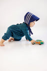 A baby in a blue bonnet and The Beaufort Bonnet Company Long Sleeve Bradford Romper in Woodlake Point Plaid with Nantucket Navy crawls on the floor, pushing a colorful toy bus.