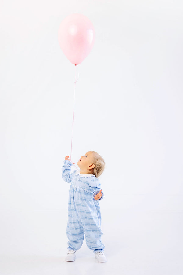 A toddler wearing The Beaufort Bonnet Company Long Sleeve Mary Dal Romper in Blue Choo Choos stands on a white background, looking up and holding a pink balloon by its string.