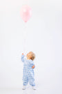 A toddler wearing The Beaufort Bonnet Company Long Sleeve Mary Dal Romper in Blue Choo Choos stands on a white background, looking up and holding a pink balloon by its string.