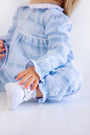 A young child wearing The Beaufort Bonnet Company’s Long Sleeve Mary Dal Romper - Blue Choo Choos paired with white sneakers is sitting on the floor. The child’s face is mostly out of frame, showing only the lower part of the face, arms, and legs.