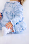 A young child wearing The Beaufort Bonnet Company’s Long Sleeve Mary Dal Romper - Blue Choo Choos paired with white sneakers is sitting on the floor. The child’s face is mostly out of frame, showing only the lower part of the face, arms, and legs.