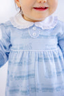 A baby wearing The Beaufort Bonnet Company’s Long Sleeve Mary Dal Romper in Blue Choo Choos, featuring a light blue design with a white lace collar and charming train patterns, smiles and shows small teeth.