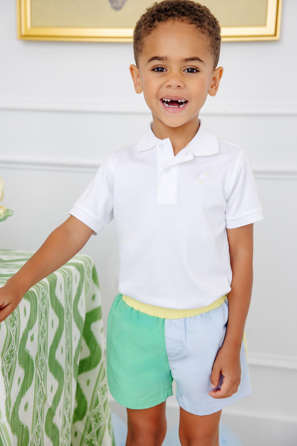 A cheerful young boy smiles while wearing the Prim & Proper Polo & Onesie in Worth Avenue White with Worth Avenue White Stork by The Beaufort Bonnet Company, paired with pastel green and blue shorts, standing indoors beside a green patterned tablecloth.