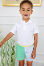 A cheerful young boy smiles while wearing the Prim & Proper Polo & Onesie in Worth Avenue White with Worth Avenue White Stork by The Beaufort Bonnet Company, paired with pastel green and blue shorts, standing indoors beside a green patterned tablecloth.