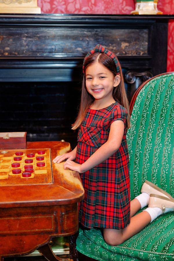 A young girl wearing The Beaufort Bonnet Company’s Darcy Dress in Paces Mill Plaid, complete with charming bow details and a coordinating headband, smiles beside a vintage checkers board while seated on a green patterned chair in an elegant red-walled room with a dark fireplace in the background.
