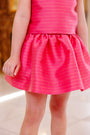 A young child is shown indoors, with only their torso and legs visible, wearing The Beaufort Bonnet Company Grandccasion™ McPrimm Party Skirt in the Raleigh Raspberry Metallic Stripe.