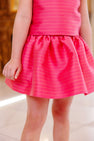 A young child is shown indoors, with only their torso and legs visible, wearing The Beaufort Bonnet Company Grandccasion™ McPrimm Party Skirt in the Raleigh Raspberry Metallic Stripe.
