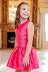Wearing The Beaufort Bonnet Company’s Grandccasion™ Tessa Top in Raleigh Raspberry Metallic Stripe, a young girl smiles and looks to the side while standing in a bright, elegant room with a fireplace and chandelier—creating the perfect scene for holiday celebrations.