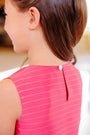 A young girl with brown hair in a ponytail is seen from the back wearing The Beaufort Bonnet Company’s Grandccasion™ Tessa Top in the Raleigh Raspberry Metallic Stripe, a sleeveless pink top featuring a small white button fastening at the neckline.