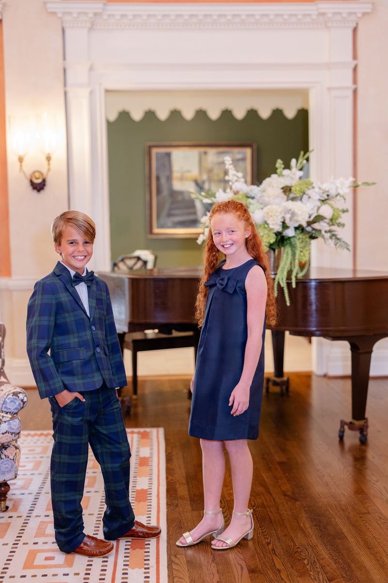 Two smiling children, a boy wearing The Beaufort Bonnet Company’s Baylor Bowtie - Berwick Black Watch plaid suit and bow tie and a girl in a navy dress and sandals, stand on a wooden floor in an elegant room with a grand floral arrangement and a piano in the background.