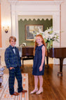 Two smiling children, a boy wearing The Beaufort Bonnet Company’s Baylor Bowtie - Berwick Black Watch plaid suit and bow tie and a girl in a navy dress and sandals, stand on a wooden floor in an elegant room with a grand floral arrangement and a piano in the background.
