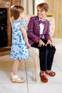 A young girl in a Beale Street Blue Grandccasion™ Delaney Dinner Dress – Bagley Bows by The Beaufort Bonnet Company chats with a boy in a red plaid blazer and bow tie, seated on a white chair. Both look joyful and beautifully dressed for a formal occasion—perfect attire for a holiday party or special event.